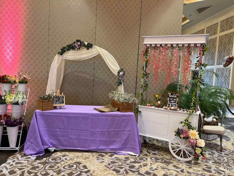 Wedding registration area with a draped table, floral arch, and a decorative cart with flowers and signs inspired by party themes.