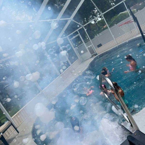 People enjoying a sunny day in a screened-in pool with a basketball hoop, a Frogger F4 Bubble Fogger Rental visible in the foreground.