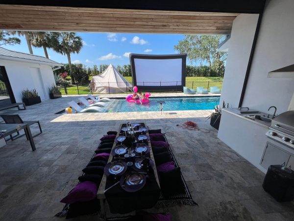 Outdoor dining setup with a long table set for a meal, overlooking a pool with a large screen and inflatable flamingo, in a luxurious backyard perfect for a Lakeland FL party.