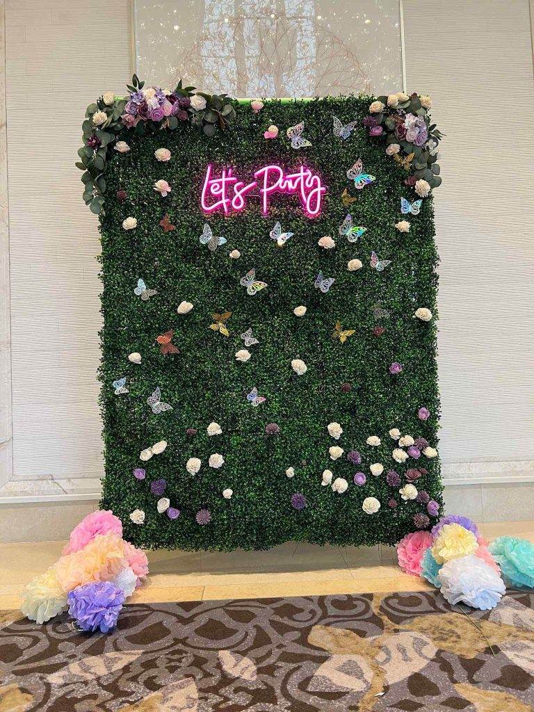 Decorative green hedge backdrop with multicolored flowers and "let's party" in neon pink signage, flanked by pastel paper pom-poms on a patterned floor, ideal for ultimate party