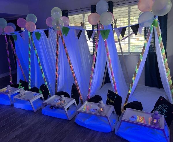Indoor sleepover setup in Tampa, FL with five tent frames, draped with white and LED-lit fabrics, surrounded by colorful balloons and personalized trays at each station.