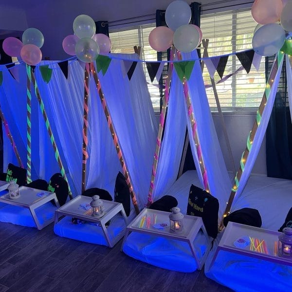 Indoor sleepover setup in Tampa, FL with five tent frames, draped with white and LED-lit fabrics, surrounded by colorful balloons and personalized trays at each station.