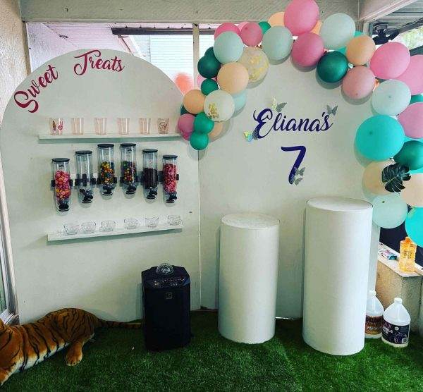 Birthday party candy station with jars of sweets, colorful balloons, and "Eliana's 7" sign, showcasing fun party themes, set up on a porch with artificial grass.
