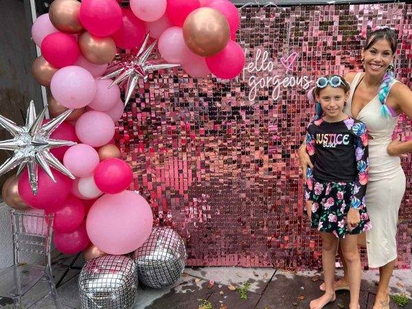 A woman and a young girl pose at a party by a pink backdrop with "hello gorgeous" sign, decorated with pink balloons and silver stars, perfect for event themes.