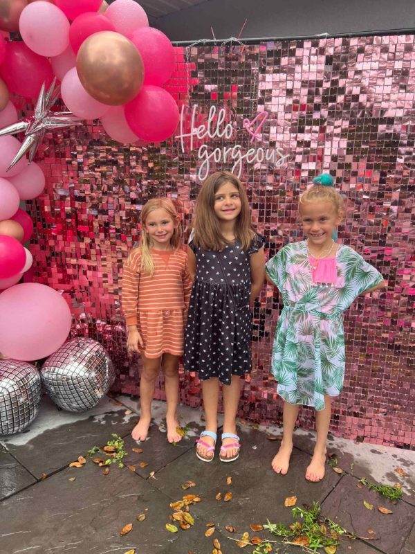 Three young girls posing in front of a pink "hello gorgeous" backdrop with balloons and pink streamers at a party themed event.