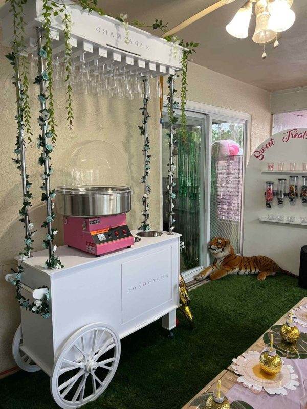 A cozy event planning space with a cotton candy machine on a decorative cart, artificial greenery, and a realistic tiger figure near a sliding glass door.