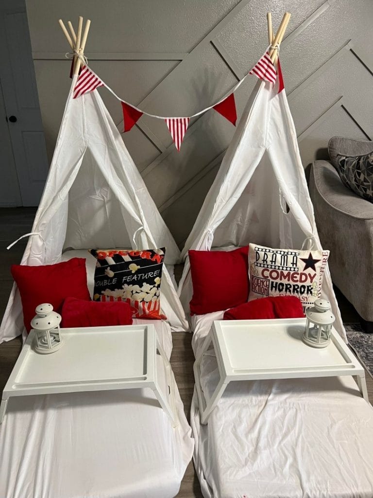 Two indoor teepee tents with white and red bedding, decorated with cinema-themed cushions and string lights, set up for a magical sleepover movie night.