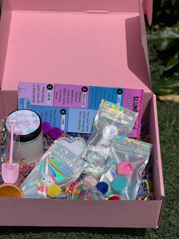 An open box filled with various craft supplies including beads, glitter, and small containers for a Slime Party Box, set on a grassy background.