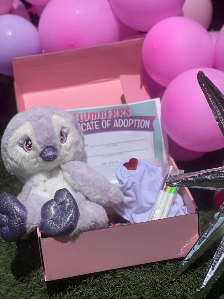 A plush toy koala sits inside a pink gift box, surrounded by pink balloons, with an adoption certificate visible, perfect for doorstep delivery.