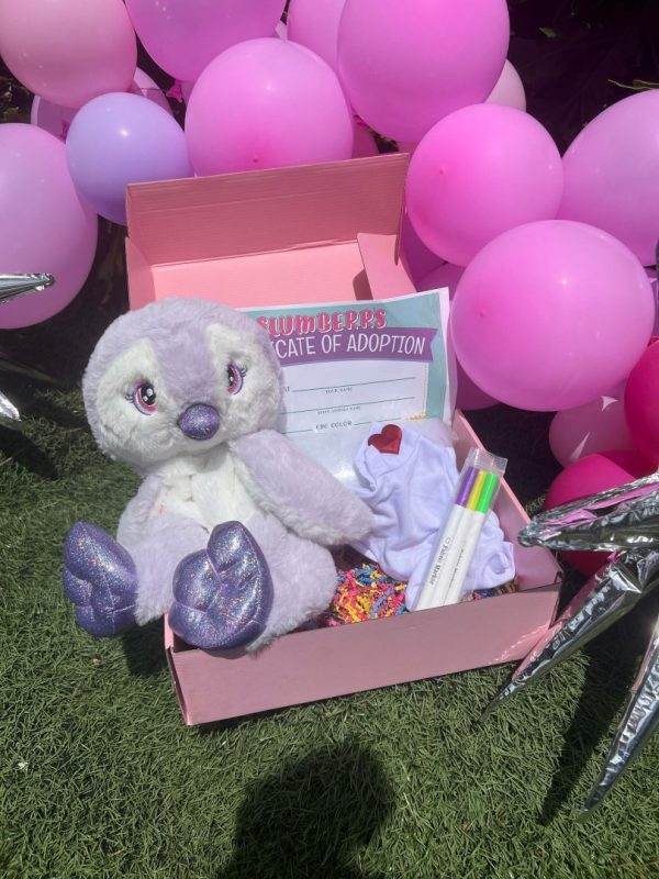 A purple plush toy with glittery accents sits inside a Slime Party Box adorned with an adoption certificate, clothes, and markers, surrounded by pink balloons and silver stars.