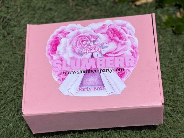 A pink Slime Party Box with a floral and bow design and the words "slime www.slumberparty.com party box" on the lid, placed on grass.