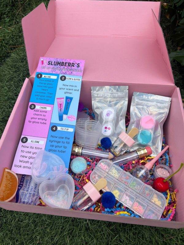An open pink DIY Lip Gloss Party Box containing various tubes, glitter packets, mixing tools, and colorful decorations on a grass background.