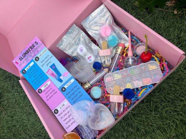 An open pink box on grass filled with various girly toys and beauty accessories, including bracelets, bottles, beads, and a DIY Lip Gloss Party Box.