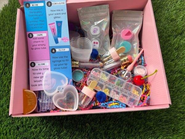 A colorful DIY Lip Gloss Party Kit with various components like glitter, scents, tubes, and a mixing guide spread out in a DIY Lip Gloss Party Box on grass.