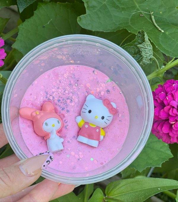 A hand holding a clear Slime Party Box of pink glittery slime with two Hello Kitty figures, surrounded by green leaves and a pink flower.