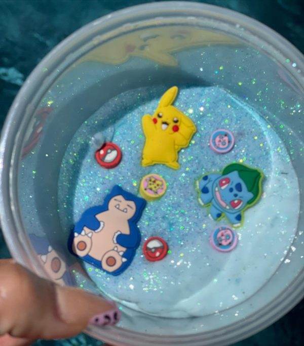 A hand holding a Slime Party Box with a glittery blue slime and various Pokémon-themed charms and badges submerged in it.