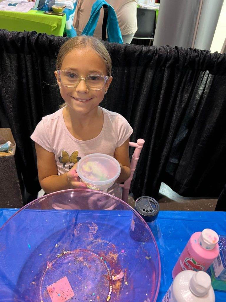 A young girl with glasses is holding a bowl of glitter slime.