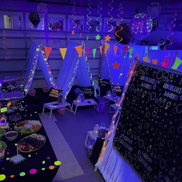 A dimly lit room decorated for the ultimate sleepover with colorful balloons, tents, a chalkboard, and a table with snacks.