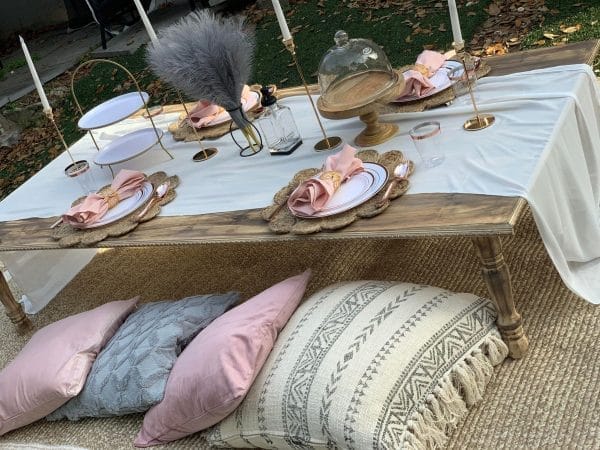 Outdoor dining setup with a rustic wooden table, white tablecloth, and elegant place settings featuring pink napkins, surrounded by cushions on the ground for the ultimate sleepover experience.