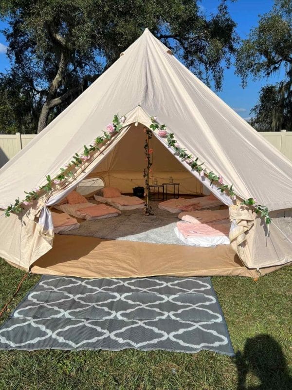 A canvas bell tent decorated with leafy garlands open to show a cozy birthday setup with rugs and cushions, set against a bright sky and greenery.
