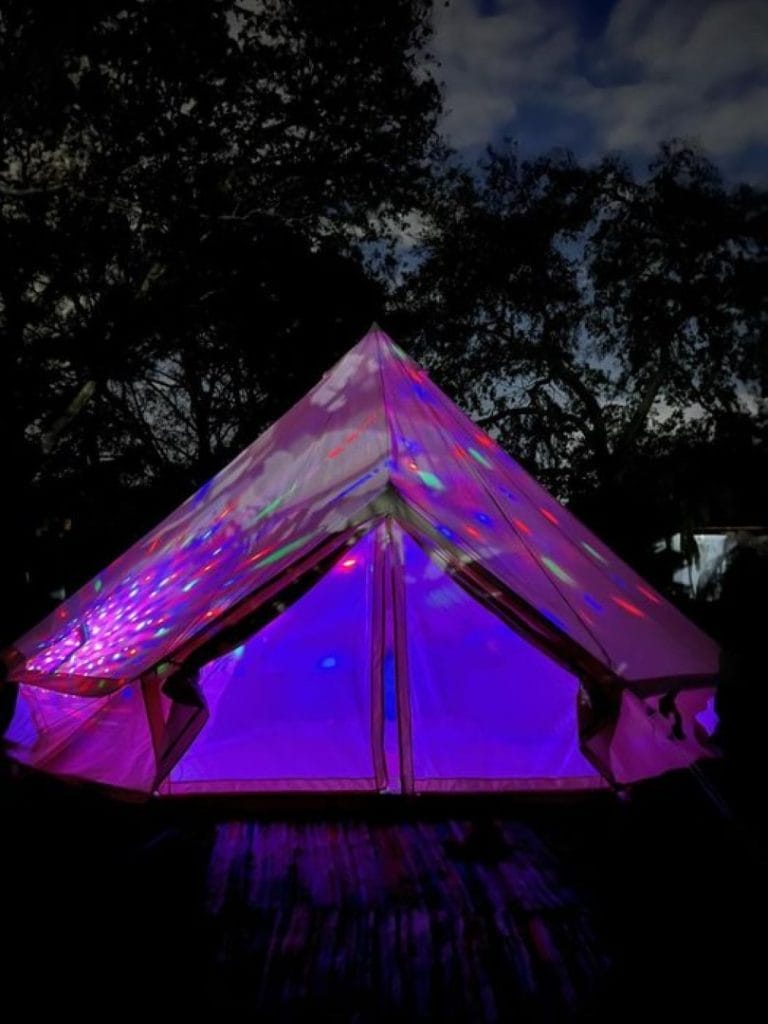 A tent illuminated with purple and multicolored lights against a dark, tree-silhouetted sky at night during an outdoor glamping birthday in Lakeland.