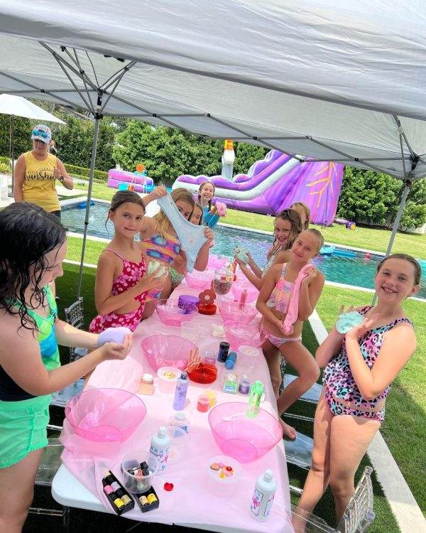 Children enjoying a craft activity with party themes at a poolside table under a canopy on a sunny day.
