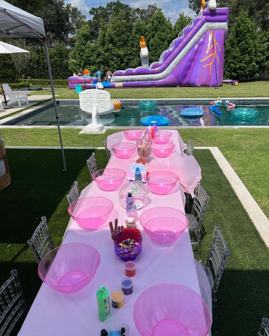 Outdoor children's party setup with a long table set with pink plates and crafts, featuring an Ultimate Sleepover theme and a large inflatable slide by the pool in the background.