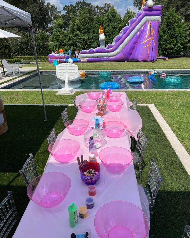 A long table set with pink plates and cups, designed for party themes, near a pool with a large inflatable slide and people in the background.