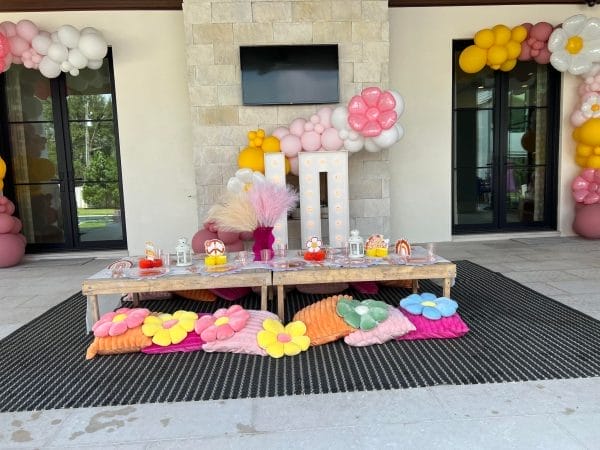 Decorative party table with colorful flower-shaped cushions, balloons, and themed decorations for an Ultimate Sleepover experience, under an outdoor covered patio.