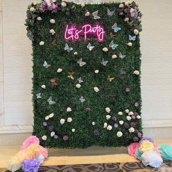 A vibrant photo booth backdrop with the words "let's party" in neon pink, surrounded by a greenery wall adorned with colorful flowers and butterflies, perfect to transform your event.