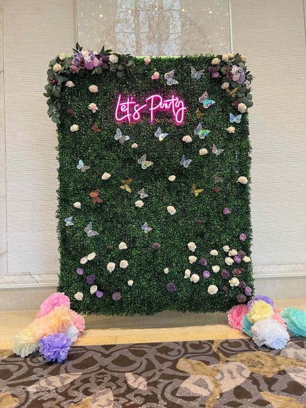 A vibrant photo booth backdrop with the words "let's party" in neon pink, surrounded by a greenery wall adorned with colorful flowers and butterflies, perfect to transform your event.