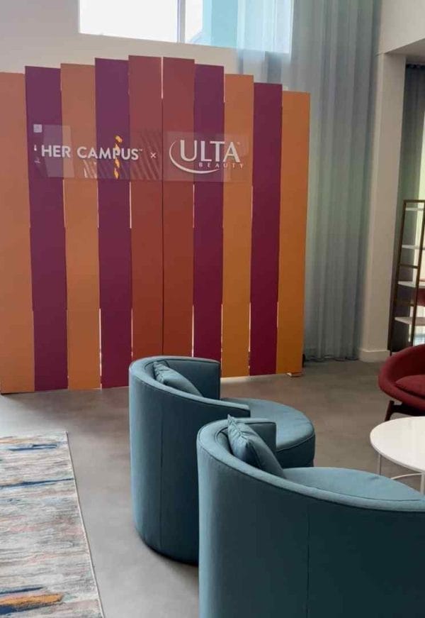 Colorful striped partition with "her campus" and "ulta" logos in a modern lounge area at a corporate event, featuring blue chairs and a decorative rug.