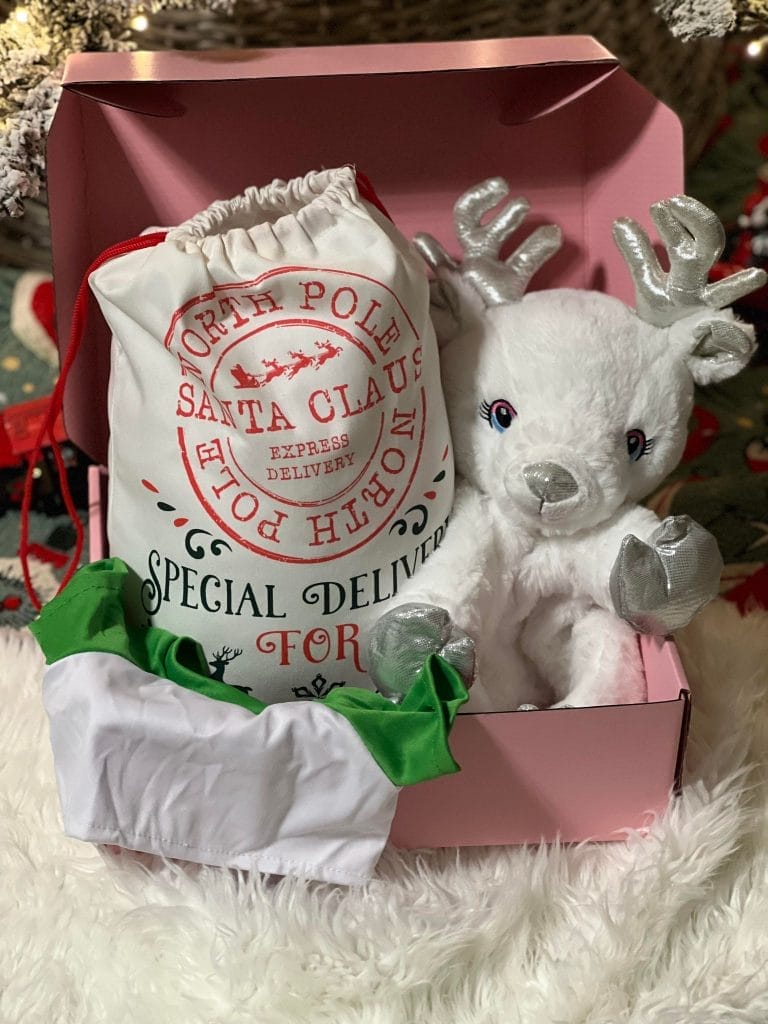 A plush white reindeer toy sits in a pink Holiday Build-A-Bear Party Box, partially wrapped in a white and red "special delivery" sack, under a Christmas tree.