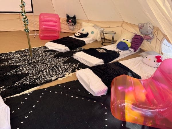 Interior of a glamping tent with four beds adorned with colorful pillows, a black and white rug, a pink chair, and party themes as decorative elements.