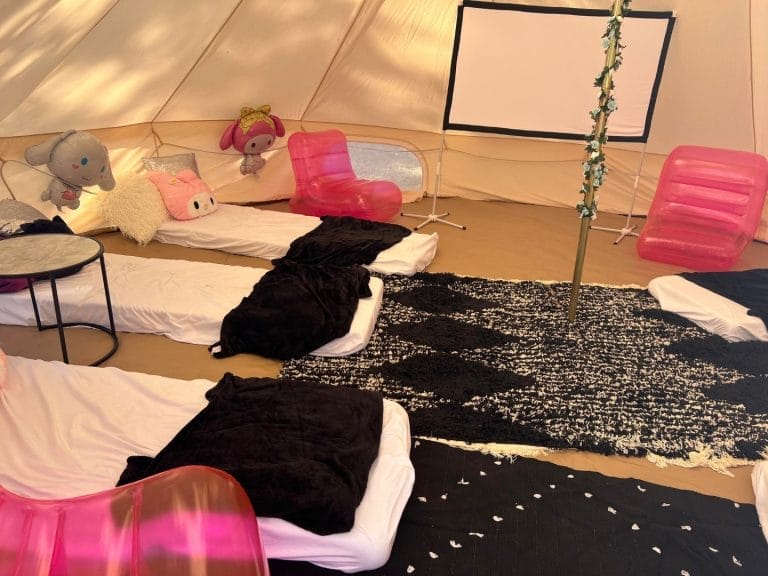 Interior of a tent with plush toys on beds, pink plastic chairs, a black rug, and scattered white petals, perfect for theme parties.