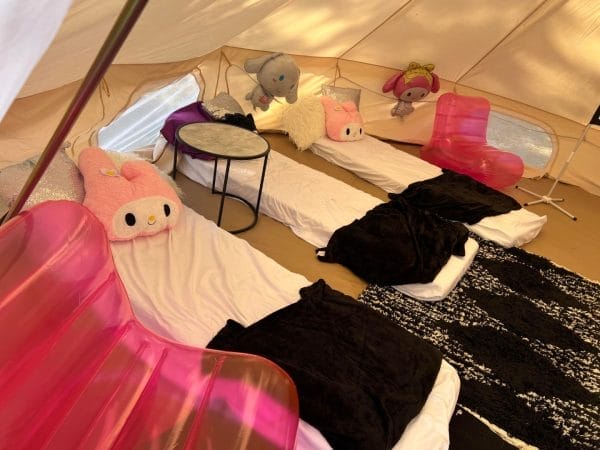 Inside a tent, there are four sleeping mats with black blankets and plush animal pillows arranged on them, surrounded by soft pink chairs and a small round table, perfectly aligned with the event planning theme.