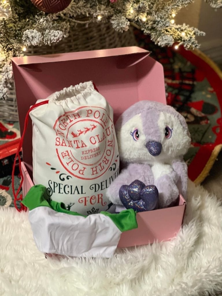 A plush toy penguin from Holiday Build-A-Bear Party Box and a cloth sack labeled "north pole santa claus special delivery" inside a pink gift box, with a Christmas tree in the background.