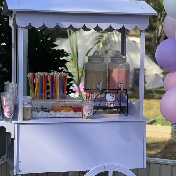 A Elegant White Snack or Candy Cart Rental decorated with Hello Kitty motifs, offering colorful straws, drinks in dispensers, and marshmallows, set against a backdrop of balloons.