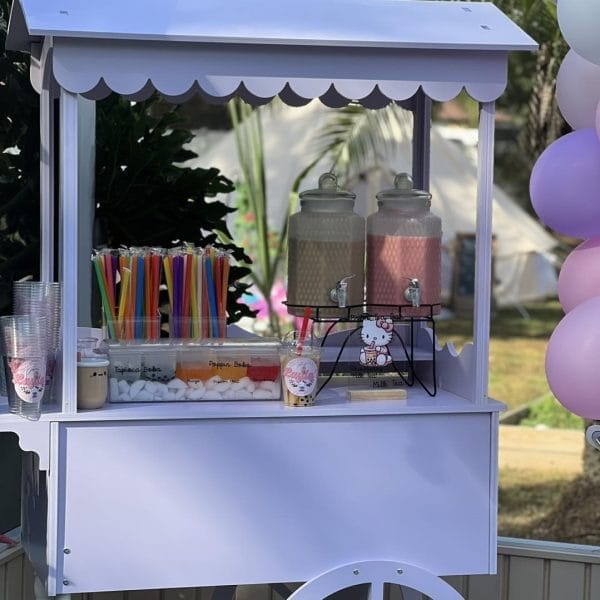 An Elegant White Snack or Candy Cart Rental with drinks in dispensers, colorful straws, and Hello Kitty decorations, surrounded by balloons.