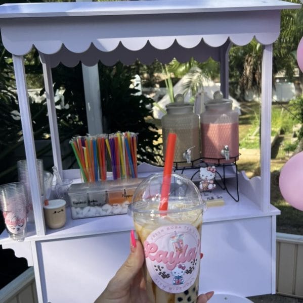 A person holding a decorated milkshake cup in front of a small outdoor Elegant White Snack or Candy Cart Rental with jars of toppings and drinks.
