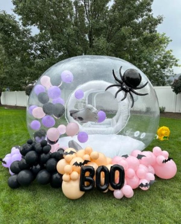 An Enchanting Bubble Balloon House Rental decorated with a black spider and balloons, surrounded by black and pink balloon clusters and a "boo" sign in a grassy yard.