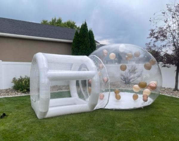 A large Enchanting Bubble Balloon House Rental with a tunnel entrance, set up in a backyard with green grass and a visible house in the background.