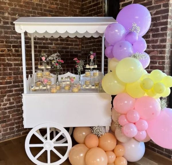 A decorative Elegant White Snack or Candy Cart rental with a canopy, adorned with a colorful cluster of balloons and displaying jars of flowers inside.