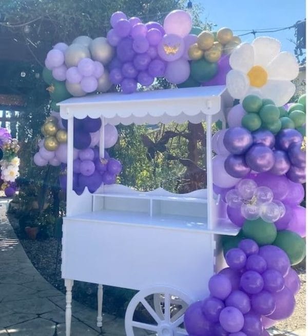 A decorative Elegant White Snack or Candy Cart adorned with clusters of purple, lavender, and white balloons, set in a sunny outdoor setting.