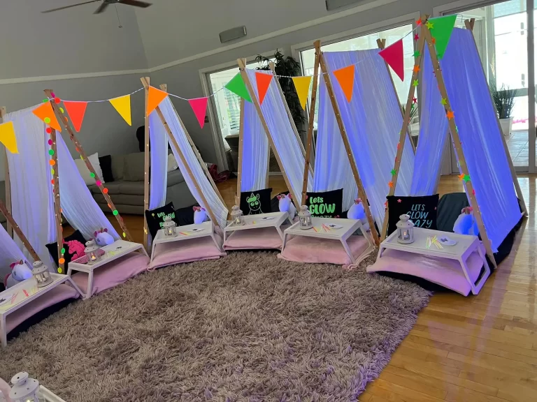 Indoor camping setup with colorful Unicorn Party Theme teepees, decorated with fairy lights and banners, arranged on a plush carpet for a children's party.