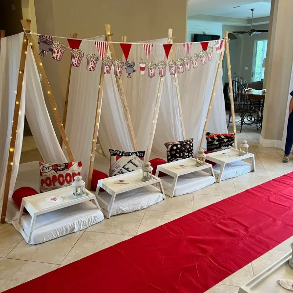 Indoor birthday party setup with a movie theme, featuring a red carpet aisle, popcorn machine, and four enchanting teepee tents with cushions.