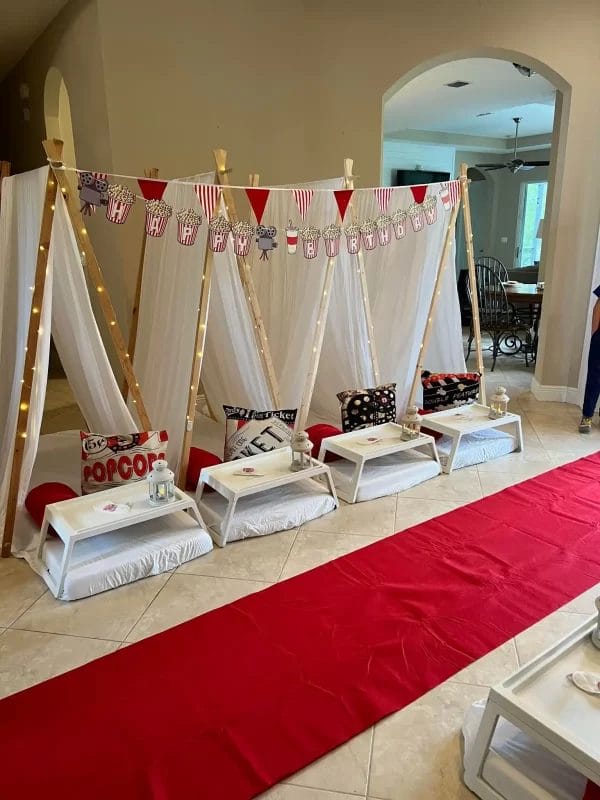 Indoor birthday party setup with a movie theme, featuring a red carpet aisle, popcorn machine, and four enchanting teepee tents with cushions.