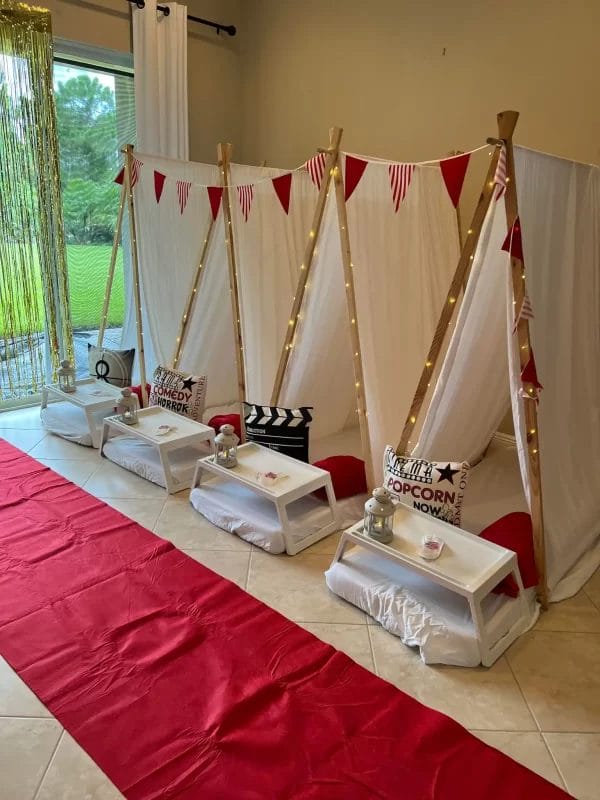 Three enchanting teepee tents with white pillows, lined up on a red carpet; each tent has a small table with popcorn and decor, under string lights.