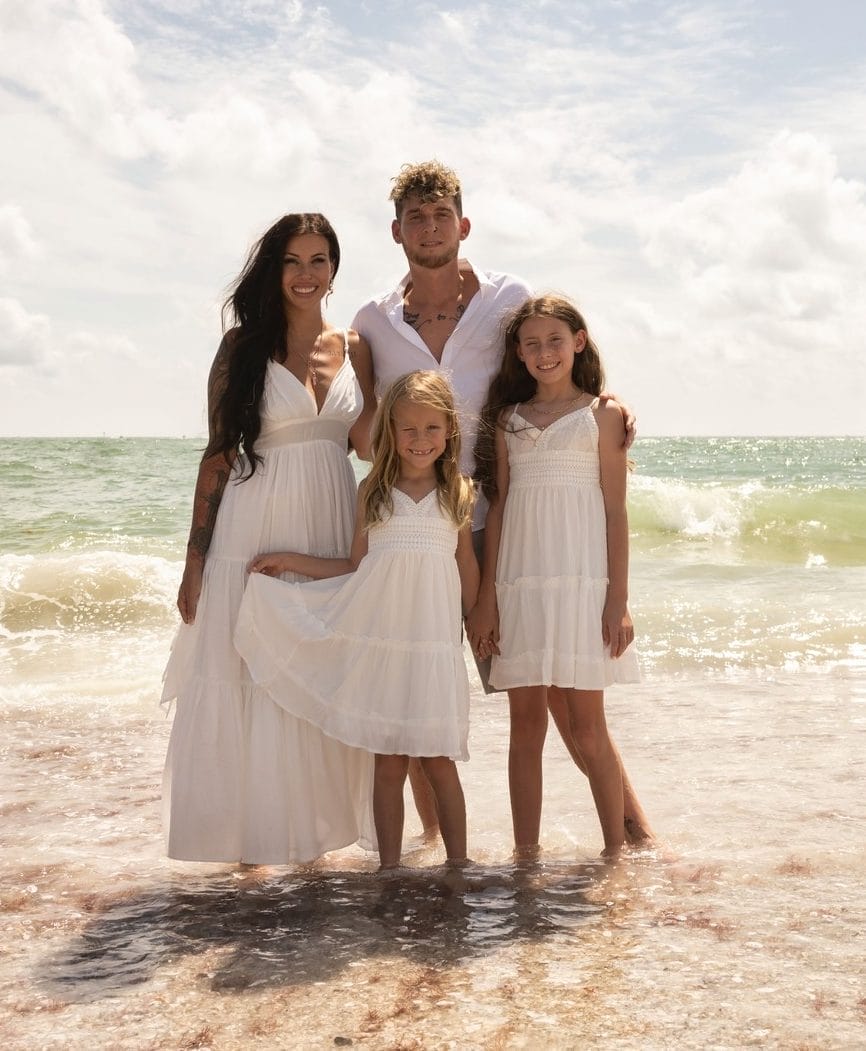 A family of four, dressed in white, standing in shallow ocean water under a sunny sky. Two adults and two children smiling towards the camera.