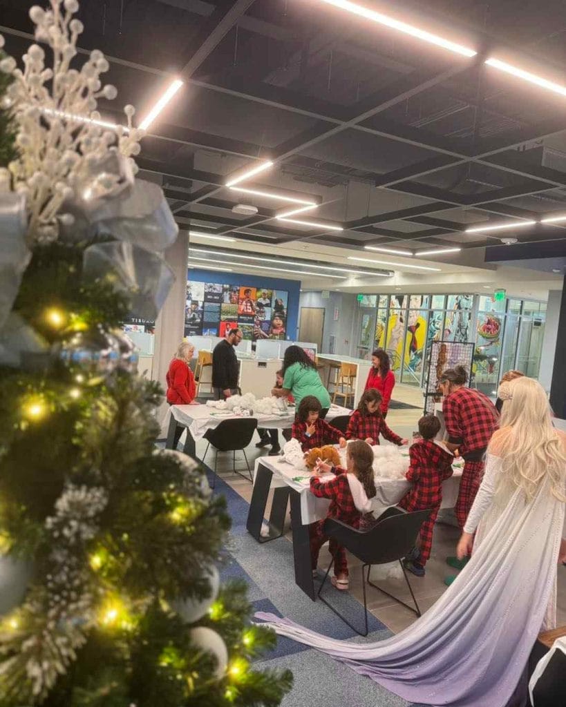 A group of children are sitting around a Christmas tree in a library, enjoying a festive party.
